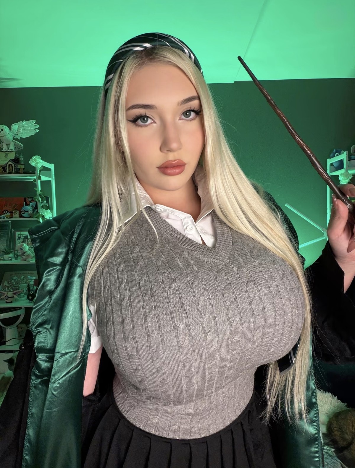 Natalia with long blonde hair, in a grey knit vest and green robe holding a wand